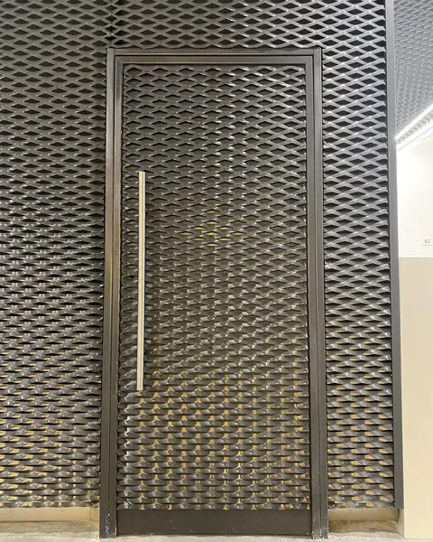 Pivot door clad in expanded metal mesh, flush with the wall, with a vertical pull handle